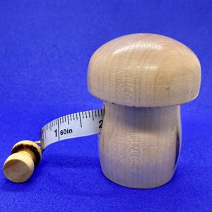 May include: A wooden mushroom-shaped measuring tape holder with a white measuring tape. The tape reads "60in" and extends around the mushroom. The mushroom is light brown and sits on a blue surface.
