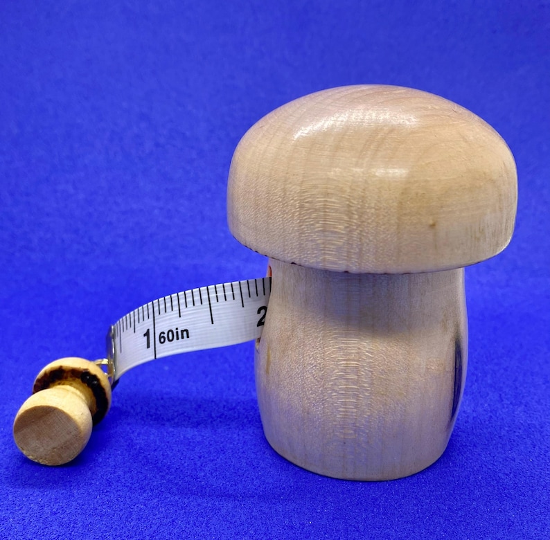 May include: A wooden mushroom-shaped measuring tape holder with a white measuring tape. The tape reads "60in" and extends around the mushroom. The mushroom is light brown and sits on a blue surface.