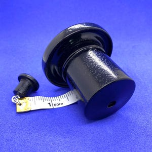 K&ouml;nnte beinhalten: Schwarzer Holz-Flaschenverschluss mit ausgestelltem Oberteil und zylindrischem K&ouml;rper. Daneben ein kleinerer Verschluss. Ein Ma&szlig;band zeigt, dass der gr&ouml;&szlig;ere Verschluss etwa 2,5 cm Durchmesser hat.