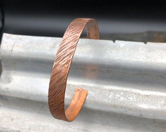 Puede incluir: Un brazalete de cobre con textura martillada. El brazalete es un diseño simple y minimalista.