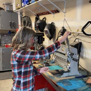 May include: A person using a metal paper trimmer to cut a piece of green paper. The trimmer is on a workbench in a garage.