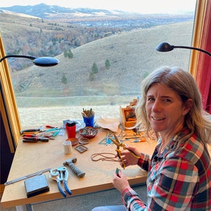 May include: A woman sits at a table with a view of a city and mountains in the background. She is working on a craft project with tools and materials spread out on the table. The tools include pliers, a hammer, a ruler, and a pair of scissors. The materials include small pieces of glass, wire, and beads.