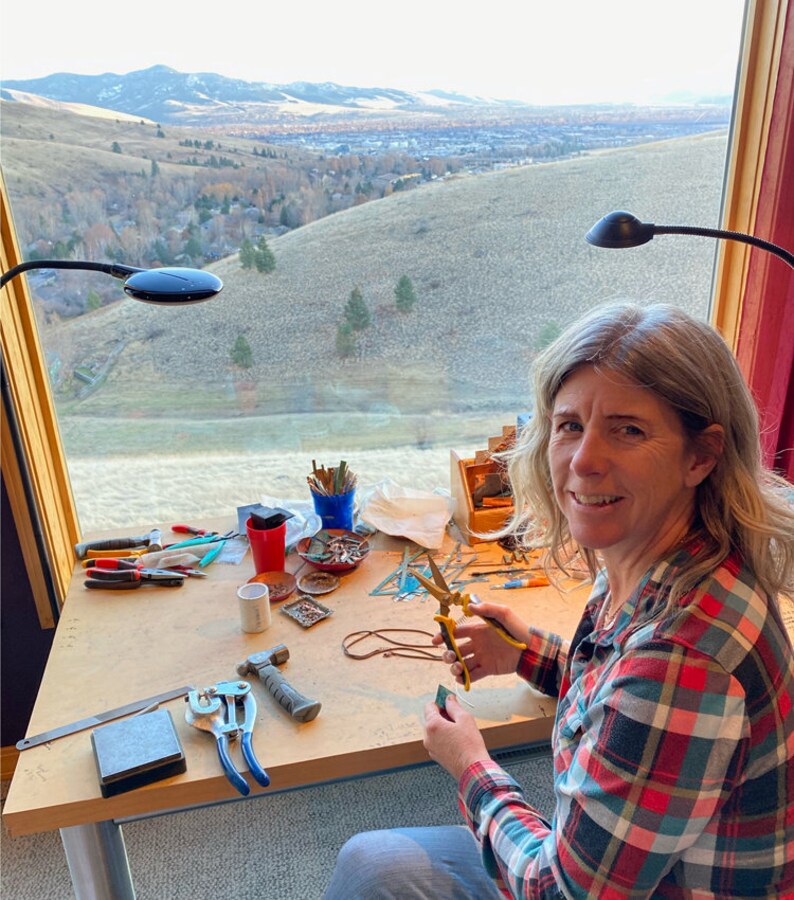 May include: A woman sits at a table with a view of a city and mountains in the background. She is working on a craft project with tools and materials spread out on the table. The tools include pliers, a hammer, a ruler, and a pair of scissors. The materials include small pieces of glass, wire, and beads.