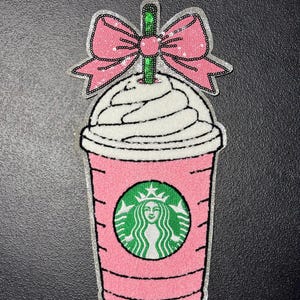 May include: Pink sequined patch in the shape of a coffee drink with a bow on top. The drink has a white whipped cream topping, a green straw, and a green circle with a woman's face in the center. The bow is pink.