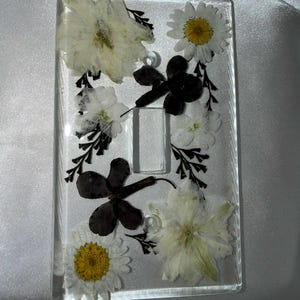 May include: A rectangular clear resin light switch cover plate featuring embedded pressed white daisy flowers, large pale petals, and dark brown foliage. The cover has a central rectangular cutout for a toggle switch and two circular screw holes.