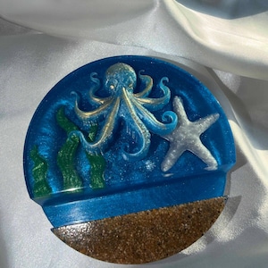 May include: A round, blue resin coaster with an ocean theme. It features a detailed octopus with gold accents, a starfish, and green seaweed. The bottom half has a sandy texture.