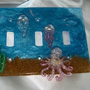 Könnte beinhalten: Eine rechteckige Abdeckung für einen Dreifach-Lichtschalter mit einer blauen Harz-Ozeanszene und Glitzer. Das Design umfasst zwei kleine silberne Quallen, einen rosa Oktopus und grüne Algenakzente auf einem strukturierten braunen Sandboden.