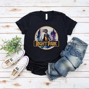 May include: A dark blue t-shirt with a watercolor illustration of a dinosaur wearing a blue shirt and standing in front of a castle. The text "Right Park" is written in a gold banner.