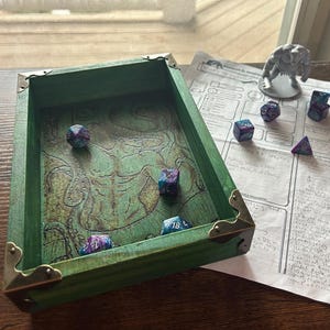 May include: A green wooden dice tray with brass corner accents, containing several multi-sided dice with blue and purple patterns. A character sheet and a small elephant figurine are also visible.