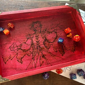 May include: A red wooden dice tray with brass corner accents. The tray features a detailed illustration of a character and several multi-sided dice in various colors. A tabletop gaming accessory.