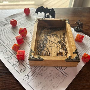 May include: A wooden dice tray with a carved image and decorative metal corners sits on a Dungeons & Dragons character sheet. Several red and orange dice are scattered around the tray, along with a small figurine and a bat-shaped cutout.