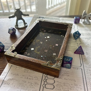 Op de afbeelding: Een houten dobbelbak met decoratieve metalen hoekaccenten. De bak is omringd door diverse polyhedrale dobbelstenen in blauw, paars en wit. Een beeldje en een spelvel zijn ook aanwezig.