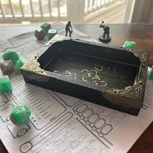 May include: A black wooden dice tray with ornate gold corner accents, surrounded by green and red dice. Two small figurines and a character sheet are also present, suggesting a tabletop role-playing game setting.