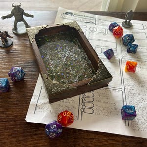 May include: A wooden dice tray with ornate metal corners, filled with glitter and small plants, sits on a wooden surface. Several colorful polyhedral dice and miniature figurines are scattered around a printed game sheet, suggesting a tabletop role-playing game.