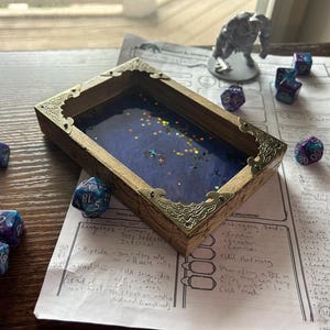 May include: A wooden dice tray with a blue interior and gold-colored decorative corners. Several multi-colored dice are scattered around the tray, along with a small figurine and a handwritten sheet of paper. The tray is designed for tabletop gaming.
