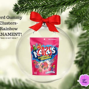May include: A clear glass ornament with a red bow, holding a pink bag of Nerd Gummy Clusters. The bag has the text "Nerd Gummy Clusters" and "Rainbow" with a colorful graphic. The ornament is hanging from a green Christmas tree branch.