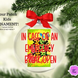 May include: Clear glass ornament with a red bow, featuring a Sour Patch Kids candy box design. The ornament has the text "IN CASE OF AN EMERGENCY BREAK OPEN" in red. The background includes a Christmas tree branch.