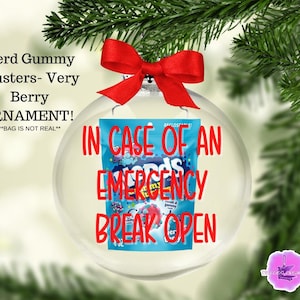 May include: A clear Christmas ornament with a red bow, containing a blue bag of Nerd Gummy Clusters. The ornament has red text that reads "IN CASE OF AN EMERGENCY BREAK OPEN". The text "Nerd Gummy Clusters- Very Berry ORNAMENT!" is also visible.