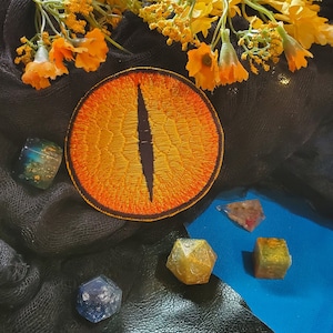 May include: A close-up of a bright orange and black embroidered eye patch with a textured surface. The eye patch is on a black fabric background with yellow flowers.