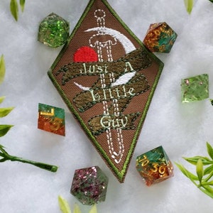 Puede incluir: Un parche de forma de diamante marrón con bordados verdes y blancos. El parche presenta una espada, una luna creciente y el texto "Just a little guy". El parche está rodeado de varios dados verdes, dorados y rojos.