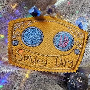 May include: A yellow embroidered patch with two blue and red circles and the text "Smiley Day".
