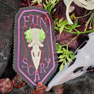 Puede incluir: Un parche bordado negro y morado con un diseño de calavera y huesos cruzados. La calavera es blanca con ojos verdes y el texto "FUN SCARY" está bordado en morado.