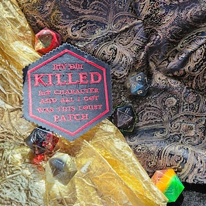 May include: A black and red embroidered patch with the text "My DM Killed My Character and all I got was this lousy patch". The patch is surrounded by several colourful dice.
