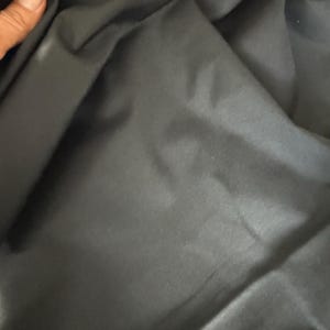 Puede incluir: Primer plano de tela negra con pliegues y arrugas visibles. La tela parece lisa, posiblemente de material sintético. La iluminación crea reflejos y sombras, enfatizando la textura y la profundidad del material.