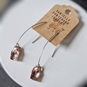 May include: A pair of copper earrings with a hammered texture, hanging from silver hoops. The earrings are displayed on a white surface with a brown card that reads "Danielle Robichaud Jewelry".