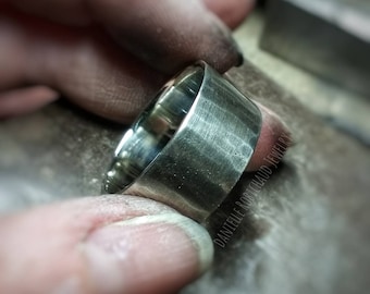 May include: A close-up of a silver ring being worked on by a jeweler. The ring is being polished and has a brushed finish. The text "DANTELLE ROBICHAUD JEWELRY" is visible on the ring.
