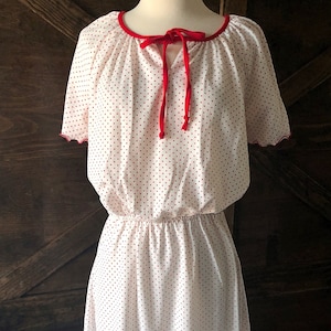 May include: A white dress with red polka dots and a red ribbon tie at the neckline. The dress has short sleeves with scalloped edges.