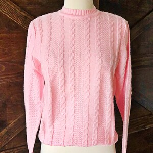 Op de afbeelding: Een roze kabel gebreide trui met lange mouwen en een ronde hals.