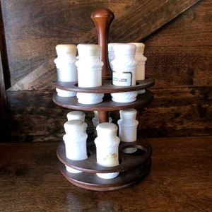 May include: A wooden spice rack with a rotating base and a handle. The rack holds 12 small white glass jars with cork tops. The jars are labeled with gold lettering. The rack is a dark brown color.