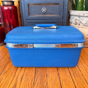 May include: A vintage blue hard-sided travel case with a silver trim and handle. The case has two silver latches on the front. The case is sitting on a wooden surface.