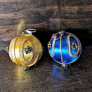Pode incluir: Dois enfeites de Natal, um dourado e outro azul, com detalhes e contas douradas. Os enfeites são feitos de tecido e têm um design tradicional.