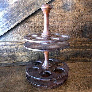 May include: A two-tiered wooden spice rack with a central post and six holes on each tier. The rack is made of dark brown wood and has a rustic appearance.