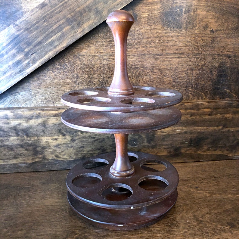 May include: A two-tiered wooden spice rack with a central post and six holes on each tier. The rack is made of dark brown wood and has a rustic appearance.