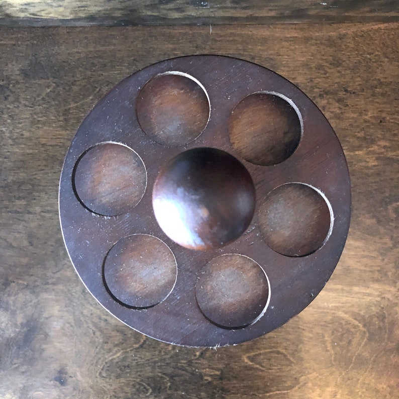 May include: A wooden egg holder with six individual compartments and a central knob. The holder is dark brown and has a rustic appearance.
