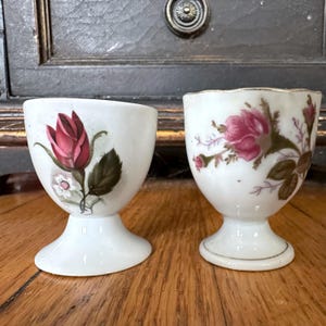 Może przedstawiać: Dwa antyczne białe porcelanowe kieliszki do jajek. Jeden kubek przedstawia czerwoną różę z zielonymi liśćmi i małym białym kwiatem. Drugi kubek ma różowy wzór róży ze złotym wykończeniem. Oba kubki mają podstawę i są wystawione na drewnianej powierzchni.