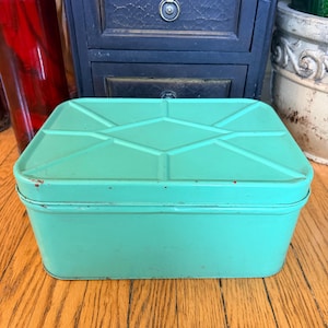Può includere: Una scatola rettangolare in metallo color verde menta con un coperchio caratterizzato da un motivo geometrico in rilievo a forma di rombo e linee. Il contenitore ha angoli arrotondati e mostra lievi segni di usura lungo i bordi del coperchio.