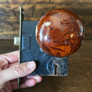 Peut inclure: Une poignée de porte en céramique marron et noire avec un motif marbré. La poignée est fixée à une plaque de porte en métal noir.