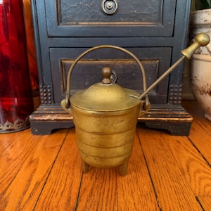 Puede incluir: Una pequeña olla de latón antigua con tapa y asa. La olla tiene un cuerpo redondo con estrías horizontales y tres patas pequeñas. Una cuchara de latón a juego descansa en el interior. La olla probablemente se usa para guardar especias u otros objetos pequeños.