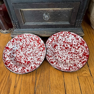 May include: Two enamelware bowls with a red and white speckled pattern. The bowls have a black rim and are set on a wooden surface. The bowls are a classic kitchenware item.
