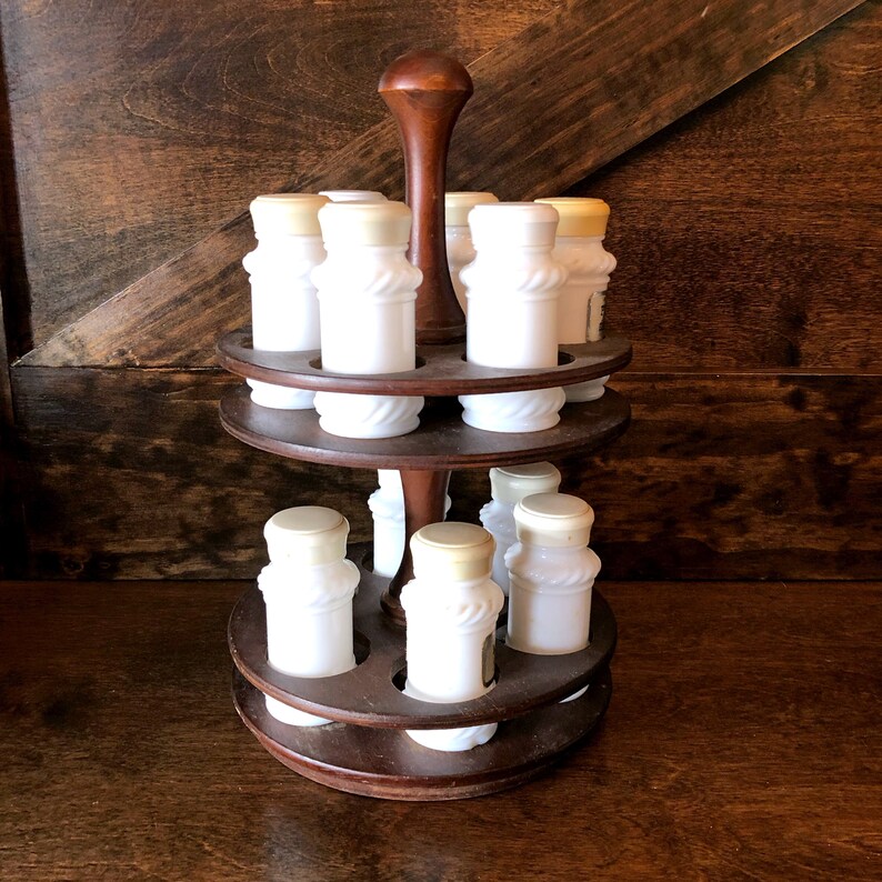May include: A wooden spice rack with two tiers holds 10 white milk glass spice jars with cork tops. The rack is dark brown wood and has a round base.
