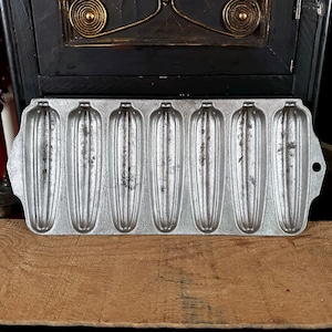 May include: A vintage cast iron cornbread pan with eight individual moulds. The pan is made of light grey metal and has a slightly textured surface.
