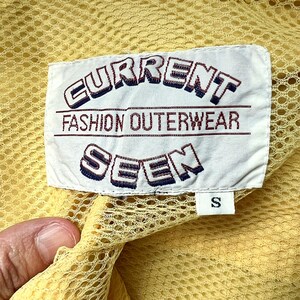 May include: A white tag with the text "CURRENT FASHION OUTERWEAR SEVEN" in a blue and red font. The tag is attached to a yellow mesh fabric.