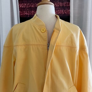 May include: A yellow bomber jacket with a zipper closure and a single button at the top. The jacket has a ribbed collar and a smooth, slightly textured fabric.