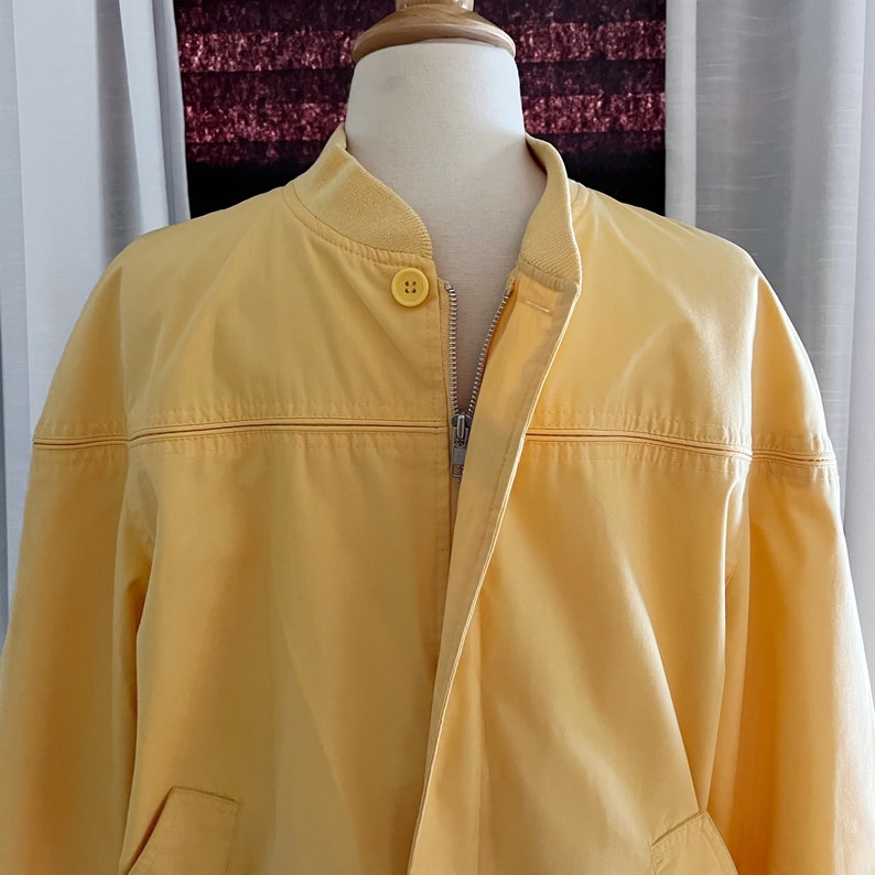 May include: A yellow bomber jacket with a zipper closure and a single button at the top. The jacket has a ribbed collar and a smooth, slightly textured fabric.