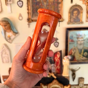 May include: An orange hourglass with a clear glass center. The hourglass is made of a marbled material and has a rounded top and base. The sand inside is white. The background includes religious art.