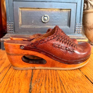 Può includere: Uno zoccolo in pelle marrone con una texture intrecciata sulla tomaia e un tacco a zeppa in legno con apertura. La scarpa presenta una punta arrotondata, un bordo in pelle intrecciata e un rivetto metallico che fissa la tomaia alla base.
